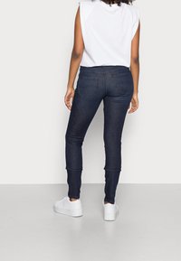 Jeans elásticos azul oscuro con corte ajustado, que presentan bolsillos traseros y una cinturilla de altura media, combinados con una blusa sin mangas blanca y zapatillas blancas.