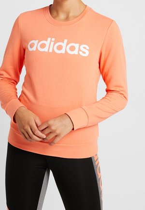 Persona que lleva una sudadera color melocotón de Adidas con logo blanco y leggings negros con marca Adidas en el lateral sobre un fondo liso.