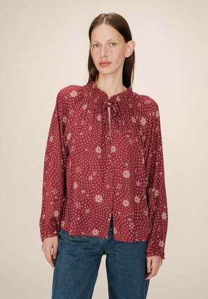 Blouse florale rouge à manches longues, décolleté rond et détail à nouer. Présente des motifs floraux blancs et bleus sur un tissu texturé.