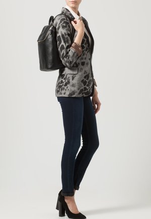 Woman wearing a gray leopard-print blazer, dark skinny jeans, black high heels, and carrying a black leather backpack over one shoulder.