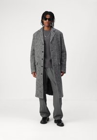 Siv houndstooth dolg plašč z črnimi gumbi, nosen preko temnega puloverja in sivih hlač; dodatki vključujejo sončna očala in srebrno verižico.