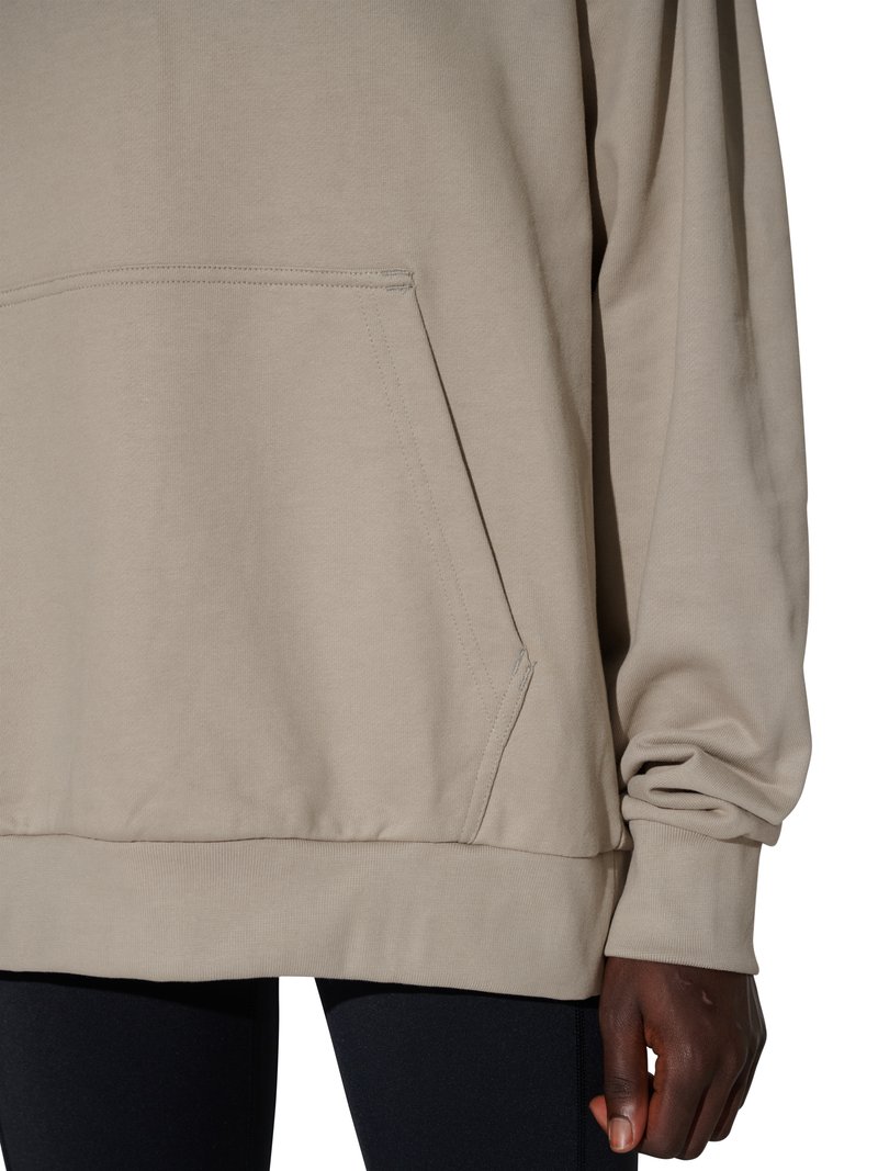 Beige sweatshirt med front-kængurulomme båret med sorte bukser, der viser højre arm og hånd mod en hvid baggrund.