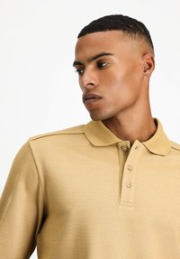 Beige Polohemd aus strukturiertem Stoff, mit einer Zwei-Knopf-Leiste und kurzen Ärmeln; mit Schulterakzenten für zusätzlichen Design-Details.