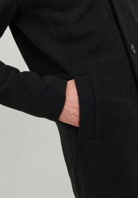 Cappotto nero realizzato in tessuto testurizzato, dotato di tasca laterale e chiusura a bottone. Vista ravvicinata che mette in evidenza il dettaglio della tasca.