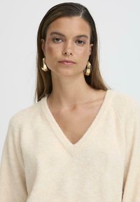 Pulover tricotat de culoare crem, cu decolteu în V adânc și mâneci raglan. O model poartă cercei mari din aur în formă de cerc, oferind un aspect texturat.