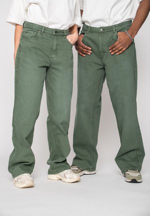 Two people wearing white shirts, green loose-fit pants, and sneakers, standing with hands in pockets against a plain background.