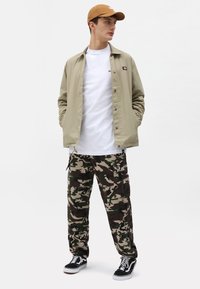 Beige jakke, hvit skjorte, kamuflasje cargo-bukser; svarte sneakers med hvite snørebånd. Brun caps. Uformell, tettsittende stil med flere lommer.