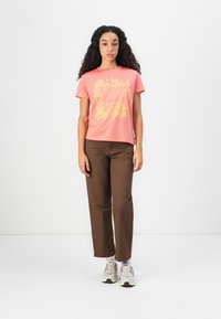 Ung kvinne med langt krøllete hår som har på seg en korallfarget T-skjorte med grafikk, brune bukser, hvite sokker og beige joggesko, stående mot en hvit bakgrunn.