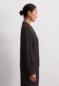Blusa lunga marrone scuro con colletto con bottoni, tessuto morbido e vestibilità rilassata. Maniche con polsini e orlo dritto. Vista laterale.
