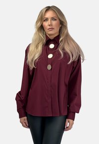 Bordowa oversize bluzka z wyraźnym kołnierzykiem, z trzema dużymi srebrnymi guzikami na przodzie oraz długimi, zwiewnymi rękawami.