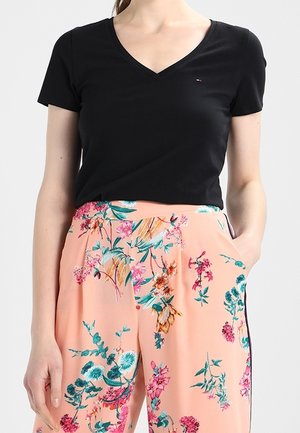 Vrouw draagt een zwart T-shirt met V-hals en korte mouwen, ingestopt in perzikkleurige broek met kleurrijk bloemenpatroon, één hand in de zak.