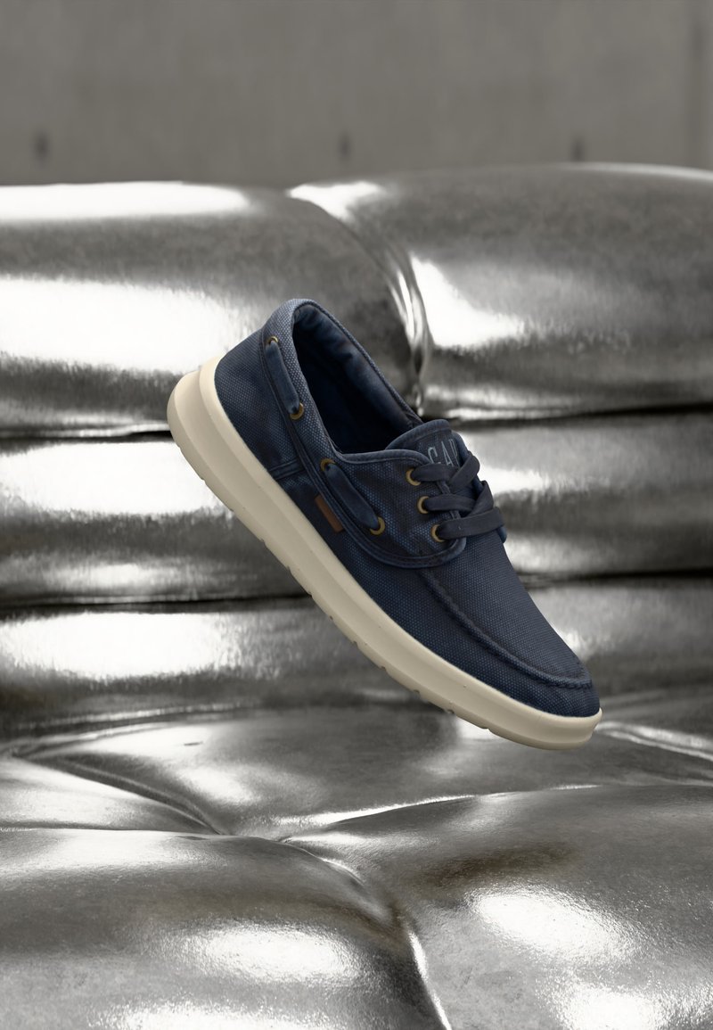 Chaussure bateau décontractée bleu marine avec semelle blanche flottant au-dessus d'un canapé en cuir argenté brillant à motifs capitonnés.