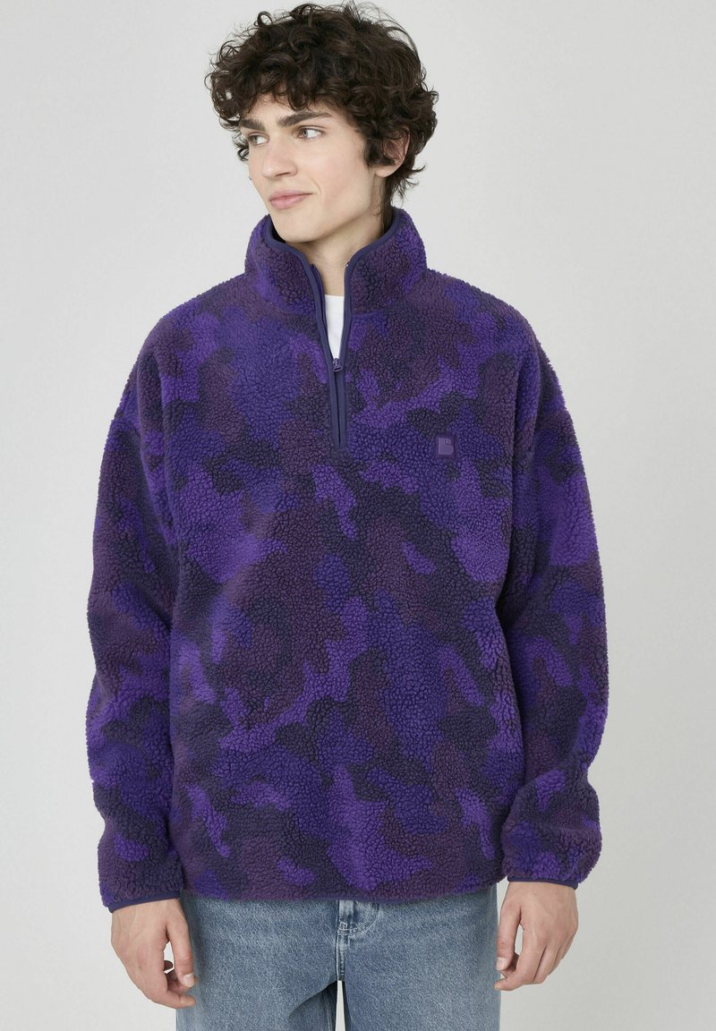 Pull en polaire violet avec col haut et zip mi-hauteur, présentant un motif camouflé dans des nuances variées de violet et de noir.