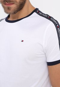 Weißes Baumwoll-T-Shirt mit navyfarbener gerippter Kragen- und Ärmelkante. Verfügt über ein kleines gesticktes Logo auf der Brust und seitliche Streifendetails.