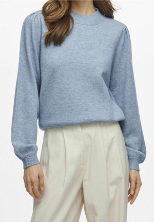 Femme portant un pull en maille bleu clair avec des manches longues bouffantes et un pantalon plissé taille haute crème, debout devant un fond blanc.
