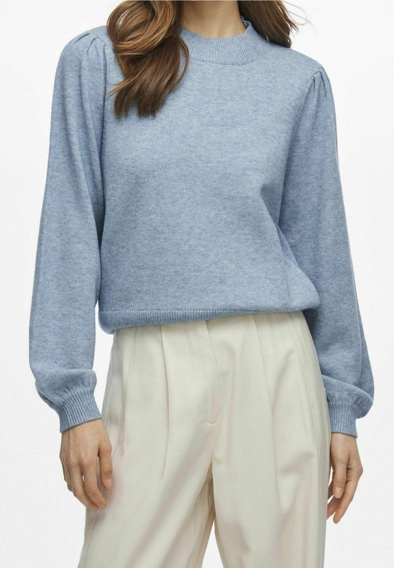 Femme portant un pull en maille bleu clair avec des manches longues bouffantes et un pantalon plissé taille haute crème, debout devant un fond blanc.