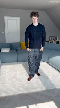 Sweater in navy with textured pattern, paired with loose dark denim pants and brown shoes. Background features a light gray corner sofa.