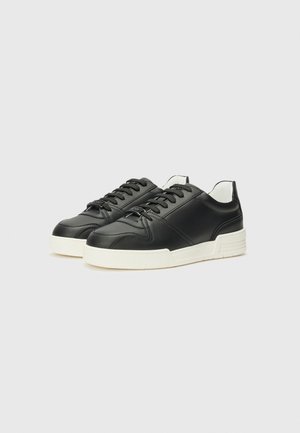 Adidași negri din piele cu o textură netedă, talpă din cauciuc alb, vârf rotund și sistem de șireturi tradițional; design minimalistic cu accente contrastante.