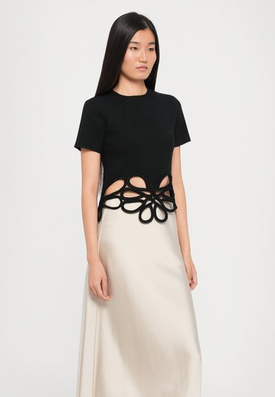 Black short-sleeve top with cut-out design, featuring a floral motif in black trim at the waist, paired with a beige flowing skirt.
