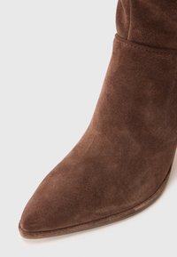 Barna suede bokacsizma hegyes orral, sima textúrával és varrás nélküli kialakítással, vékony, magasított sarkával.