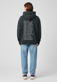 Kolen hoodie met een grote capuchon, met witte verticale tekst op de achterkant. Lichtblauwe spijkerbroek met een relaxed fit en subtiele textuur.