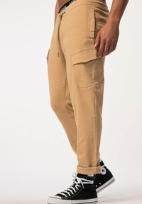 Beige Cargo-Hosen mit verstellbarem Bund, seitlichen Taschen und gesäumten Bündchen, kombiniert mit schwarzen High-Top-Sneakern. Der Stoff wirkt weich und bequem.