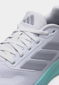 Zapatilla blanca de Adidas con cordones blancos, paneles laterales sintéticos texturizados y una suela azul clara, vista en primer plano.
