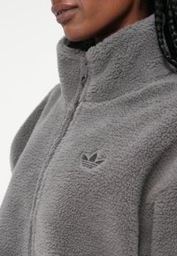 Giacca in pile grigia con colletto alto, zip e un discreto logo sul petto. Tessuto morbido e vestibilità rilassata, ideale per un abbigliamento casual.