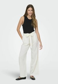 Černý žebrovaný top na ramínka v kombinaci s bílými lněnými kalhotami s vysokým pasem a uvázaným páskem. Modelka má na sobě černé baleríny. Jednoduchý, ležérní outfit.