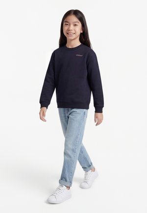 Νεαρό άτομο περπατά φορώντας μπλε σκούρο φούτερ με το κείμενο "Off-White", ανοιχτόχρωμο τζιν γυρισμένο στις άκρες και λευκά αθλητικά παπούτσια.