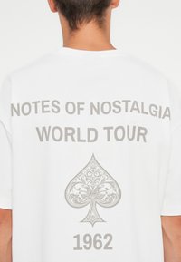 Witte katoenen t-shirt met grijze tekst: "NOTES OF NOSTALGIA WORLD TOUR" en een decoratieve klavergrafiek met "1962" eronder.