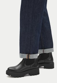 Czarne skórzane botki za kostkę z grubymi, teksturowanymi podeszwami i elastycznymi bocznymi panelami, połączone z ciemnymi dżinsami z podwiniętymi mankietami.