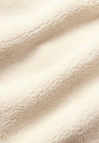 Cremefarbenes Fleece-Gewebe mit einer weichen, strukturierten Oberfläche. Verfügt über ein welliges Muster, das sein plüschiges und kuscheliges Aussehen unterstreicht.