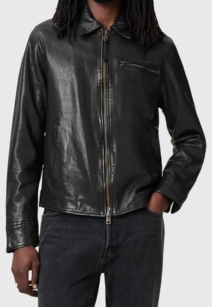 Muž v černé kožené bundě se zipem vpředu a náprsní kapsou, doplněné tmavými džínami, s rukou v kapse na jednobarevném pozadí.