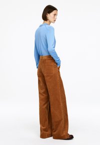 Frau in einem hellblauen Langarmshirt und hoch taillierten, weiten braunen Cordhosen, die mit den Händen in den Taschen seitlich steht.