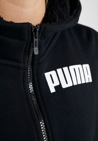 Gros plan d'une veste noire zippée avec un col en polaire et un logo Puma blanc sur la poitrine. La fermeture éclair est partiellement fermée.