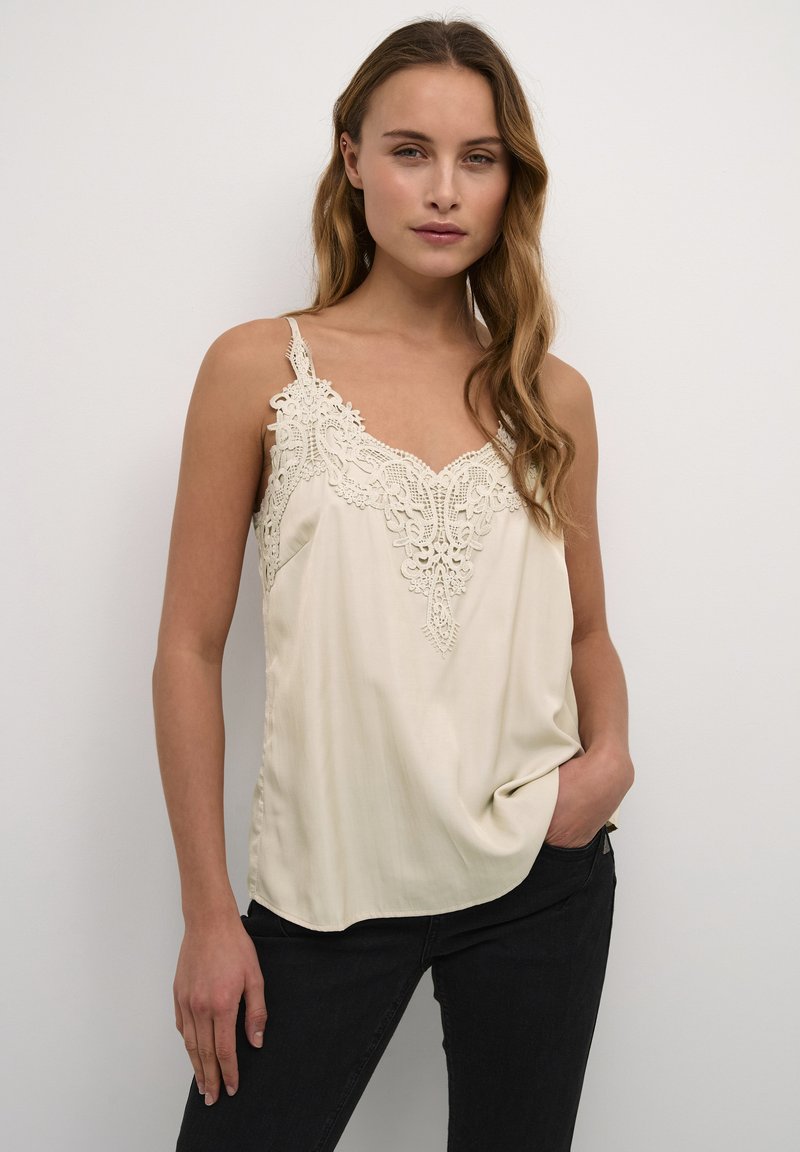 Beige silk camisole dengan detail renda di leher dan tali tipis, dipadukan dengan celana jeans hitam, dengan latar belakang netral.