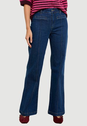 Vrouw draagt donkerblauwe rechte jeans met hoge taille en voornaaddetail, gecombineerd met bordeauxrode schoenen en een roze-zwarte gestreepte trui.