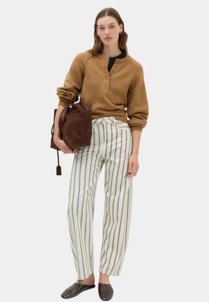 Kvinde iført en brun sweater og hvide stribede bukser holder en brun taske og står afslappet med mørke slip-on sko.