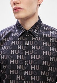 Μαύρο πουκάμισο με επαναλαμβανόμενο τύπωμα "HUG" σε διάφορους τόνους του γκρι. Σχεδίαση με κουμπιά και αιχμηρό γιακά, από υφή υφάσματος.