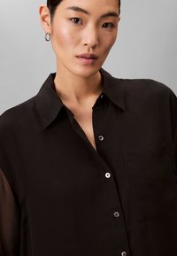 Sort gennemsigtig bluse med krave, lukning foran med knapper, en enkelt lomme til venstre og sølvfarvede knapper.