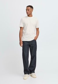 Camiseta blanca de manga corta combinada con jeans rectos de color azul oscuro y zapatillas de color claro, con un fondo liso claro.