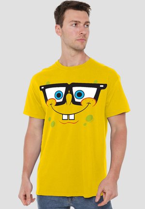 Gelbes Baumwoll-T-Shirt mit einem großen bedruckten Grafikdesign eines lächelnden Charakters mit Brille, blauen Augen und grünen Akzentpunkten.