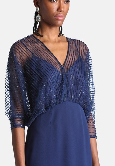 Femme portant une robe bleu marine à col en V avec des manches transparentes superposées ornées de perles et de grandes boucles d’oreilles rectangulaires en cristal.