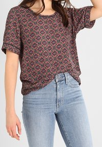 Frau in einem kurzärmeligen gemusterten Oberteil mit geometrischem Blumenmuster, das in eine hochgeschnittene hellblaue Jeans gesteckt ist.