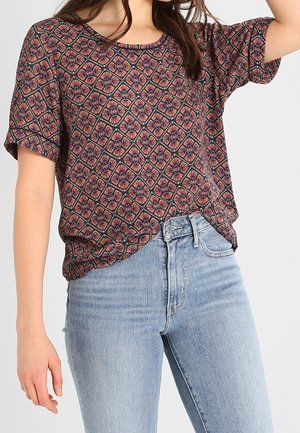 Woman wearing short-sleeve patterned top with geometric floral design tucked into high-waisted light blue jeans.