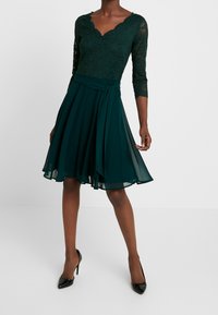 Spets- och chiffongklänning i mörkgrön med trekvartslång ärm, v-ringad halsringning och figurnära liv som övergår till en A-linjeformad kjol.
