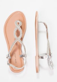 Hopeasävyiset litteät sandaalit, joissa on koristeellinen, toisiinsa punottu hihnasuunnittelu. Ominaisuuksiin kuuluu sileä vaalea ruskea pohja ja teksturoitu kumipohja, joka tarjoaa pitoa.