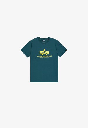 T-shirt in cotone teal con un logo giallo ben visibile "Alpha Industries" con accenti di ali sul davanti. Collo a giroclassico e maniche corte.