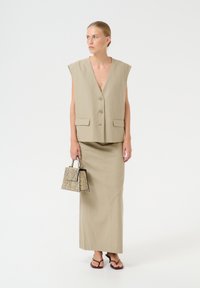 Bežs lins vestes ar dziļu V veida kaklu un priekšējiem pogām, apvienotā ar saderīgu garu svārku. Tur mazu rokassomu ar čūsku ādas rakstu.